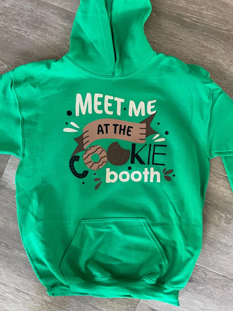 May include: Green hooded sweatshirt with the text "Meet me at the cookie booth" in white and brown lettering. A brown and white cookie graphic is included.