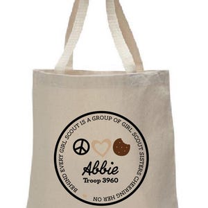 May include: Beige canvas tote bag with cream-colored handles. The bag features a circular design with the text "EVERY GIRL SCOUT IS A GROUP OF GIRL SCOUT SISTERS CHEERING HER ON." The design includes a peace sign, heart, and cookie. The name "Abbie" and "Troop 3960" are also displayed.