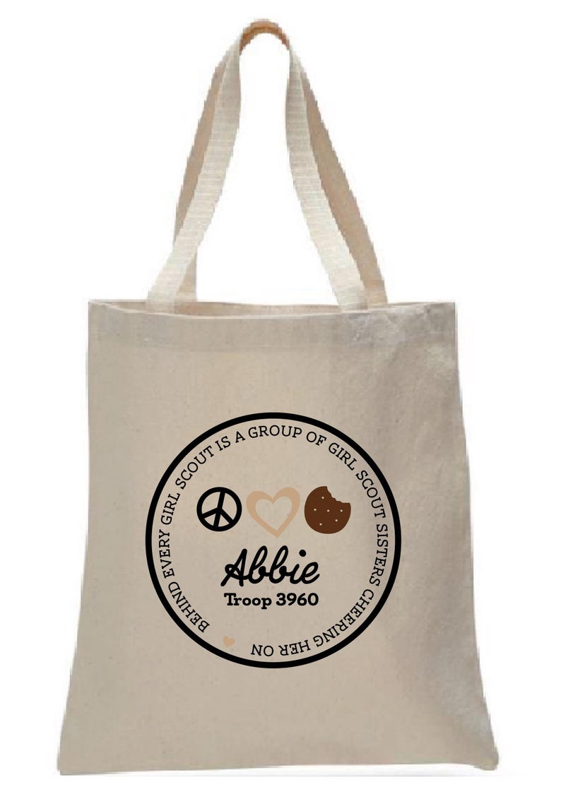 May include: Beige canvas tote bag with cream-colored handles. The bag features a circular design with the text "EVERY GIRL SCOUT IS A GROUP OF GIRL SCOUT SISTERS CHEERING HER ON." The design includes a peace sign, heart, and cookie. The name "Abbie" and "Troop 3960" are also displayed.