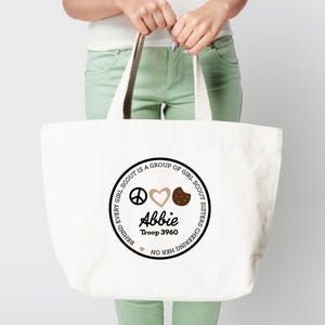 May include: A white canvas tote bag with a circular design featuring the text "BEHIND EVERY GIRL SCOUT IS A GROUP OF GIRL SCOUT SISTERS CHEERING HER ON." The design includes a peace sign, heart, cookie, the name "Abbie," and "Troop 3960."