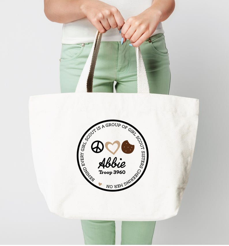 May include: A white canvas tote bag with a circular design featuring the text "BEHIND EVERY GIRL SCOUT IS A GROUP OF GIRL SCOUT SISTERS CHEERING HER ON." The design includes a peace sign, heart, cookie, the name "Abbie," and "Troop 3960."