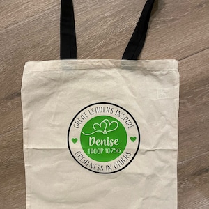 Pu&ograve; includere: Una borsa tote in tela bianca con manici neri. La borsa ha un cerchio verde con il testo "Great Leaders Inspire" e "Denise Troop 10256" in testo nero. Il cerchio contiene anche due cuori e il testo "Greatness in Others" in testo nero.
