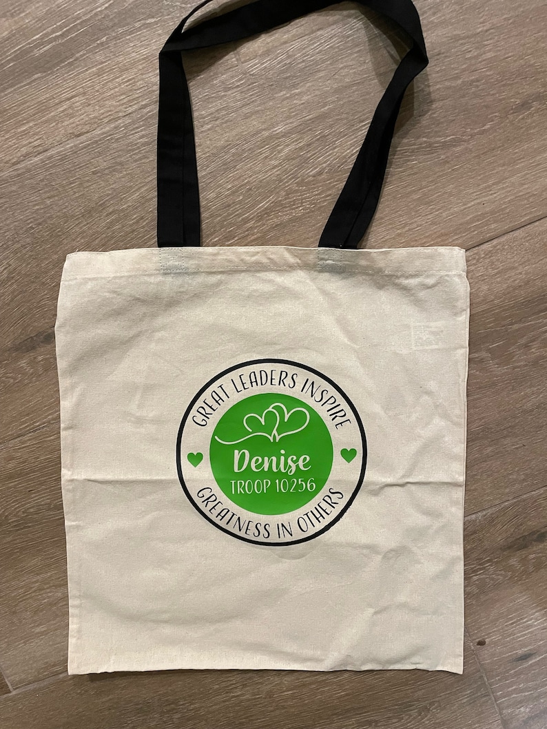 Pu&ograve; includere: Una borsa tote in tela bianca con manici neri. La borsa ha un cerchio verde con il testo "Great Leaders Inspire" e "Denise Troop 10256" in testo nero. Il cerchio contiene anche due cuori e il testo "Greatness in Others" in testo nero.