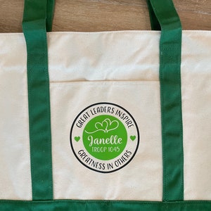 Pu&ograve; includere: Borsa a tracolla in tela verde con una tasca bianca che presenta un cerchio nero con il testo "Great Leaders Inspire" e "Janelle Troop 1045 Greatness in Others" in bianco. Il cerchio contiene due cuori verdi.