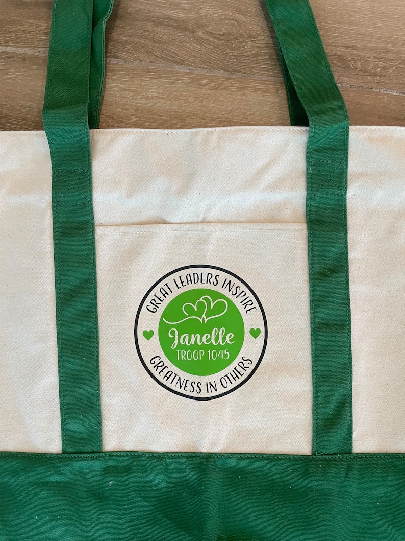 Pu&ograve; includere: Borsa a tracolla in tela verde con una tasca bianca che presenta un cerchio nero con il testo "Great Leaders Inspire" e "Janelle Troop 1045 Greatness in Others" in bianco. Il cerchio contiene due cuori verdi.