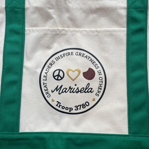 May include: Cream-colored tote bag with green straps and a front pocket. The bag features a circular emblem with the text "GREAT LEADERS INSPIRE GREATNESS IN OTHERS." The emblem includes a peace sign, a gold heart, a cookie, and the name "Marisela" and "Troop 3780."
