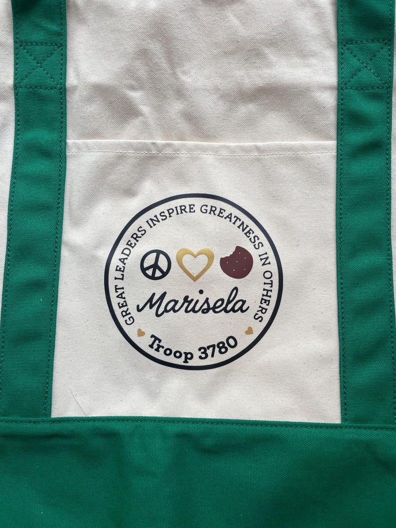 May include: Cream-colored tote bag with green straps and a front pocket. The bag features a circular emblem with the text "GREAT LEADERS INSPIRE GREATNESS IN OTHERS." The emblem includes a peace sign, a gold heart, a cookie, and the name "Marisela" and "Troop 3780."
