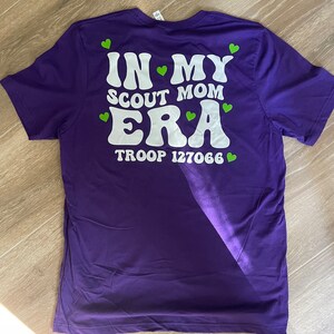 May include: Purple t-shirt with white text that reads "In My Scout Mom Era" and "Troop 127066". Green hearts are scattered around the text.