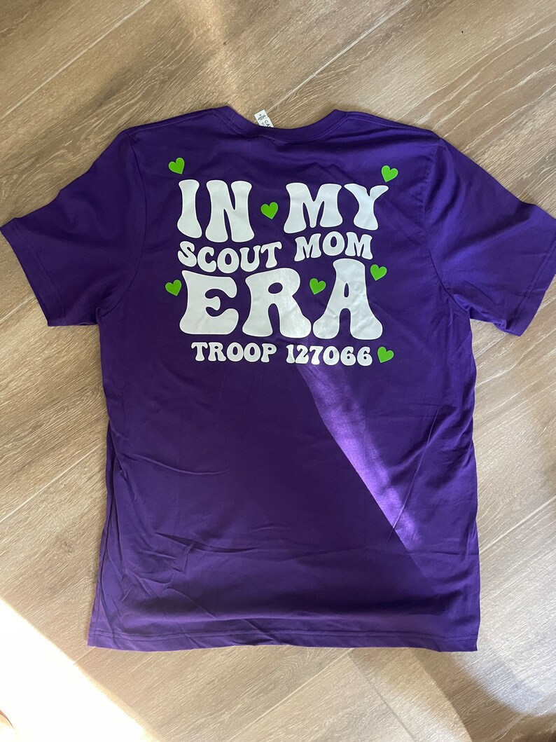 May include: Purple t-shirt with white text that reads "In My Scout Mom Era" and "Troop 127066". Green hearts are scattered around the text.