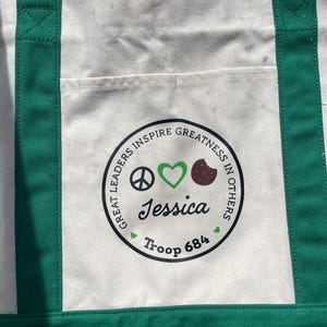 May include: Cream-colored canvas tote bag with green handles and trim. The bag features a circular emblem with the text "GREAT LEADERS INSPIRE GREATNESS IN OTHERS." The emblem includes a peace sign, a green heart, a brown cookie, and the name "Jessica" and "Troop 684."