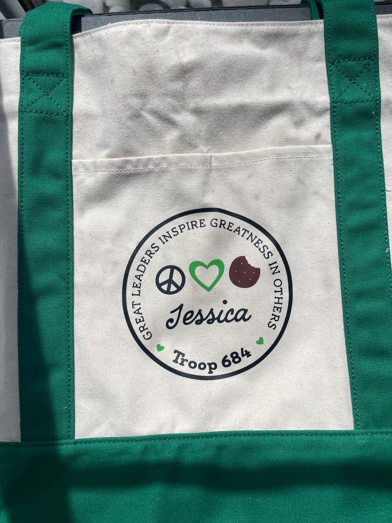 May include: Cream-colored canvas tote bag with green handles and trim. The bag features a circular emblem with the text "GREAT LEADERS INSPIRE GREATNESS IN OTHERS." The emblem includes a peace sign, a green heart, a brown cookie, and the name "Jessica" and "Troop 684."