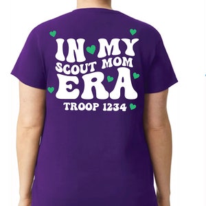 May include: Three t-shirts with the text "IN MY SCOUT MOM ERA" and "TROOP 1234" printed on the back. The shirts are green, purple, and blue.