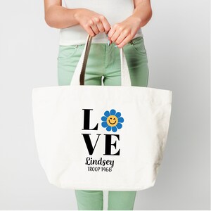 Pu&ograve; includere: Borsa tote in tela bianca con testo nero che recita "LOVE" con un fiore blu con un'emoticon gialla sorridente nella "O". Sotto il testo c'&egrave; "Lindsey TROOP 1468".