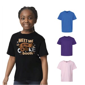Puede incluir: Una persona joven lleva una camiseta negra con el texto "MEET ME AT THE COOKIE booth" en blanco y marr&oacute;n. Tambi&eacute;n se muestran tres camisetas lisas en azul, morado y rosa.