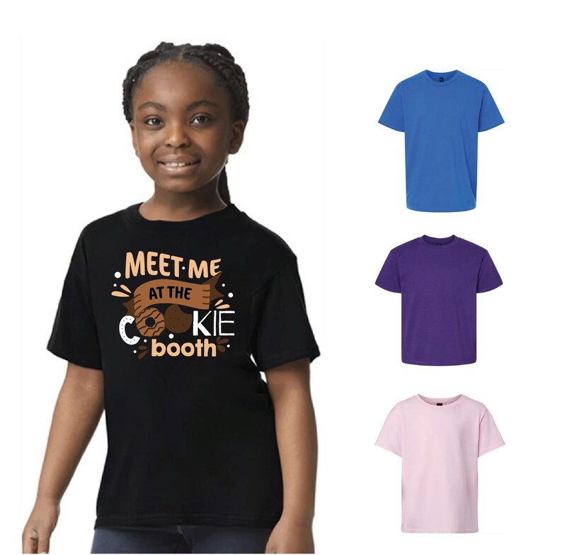 Puede incluir: Una persona joven lleva una camiseta negra con el texto "MEET ME AT THE COOKIE booth" en blanco y marr&oacute;n. Tambi&eacute;n se muestran tres camisetas lisas en azul, morado y rosa.