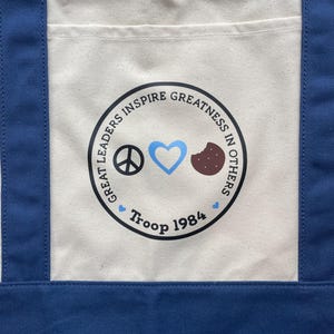 May include: A blue and cream tote bag with a circular graphic. The graphic includes a peace sign, a blue heart, and a cookie. The text reads "Great Leaders Inspire Greatness in Others" and "Troop 1984".