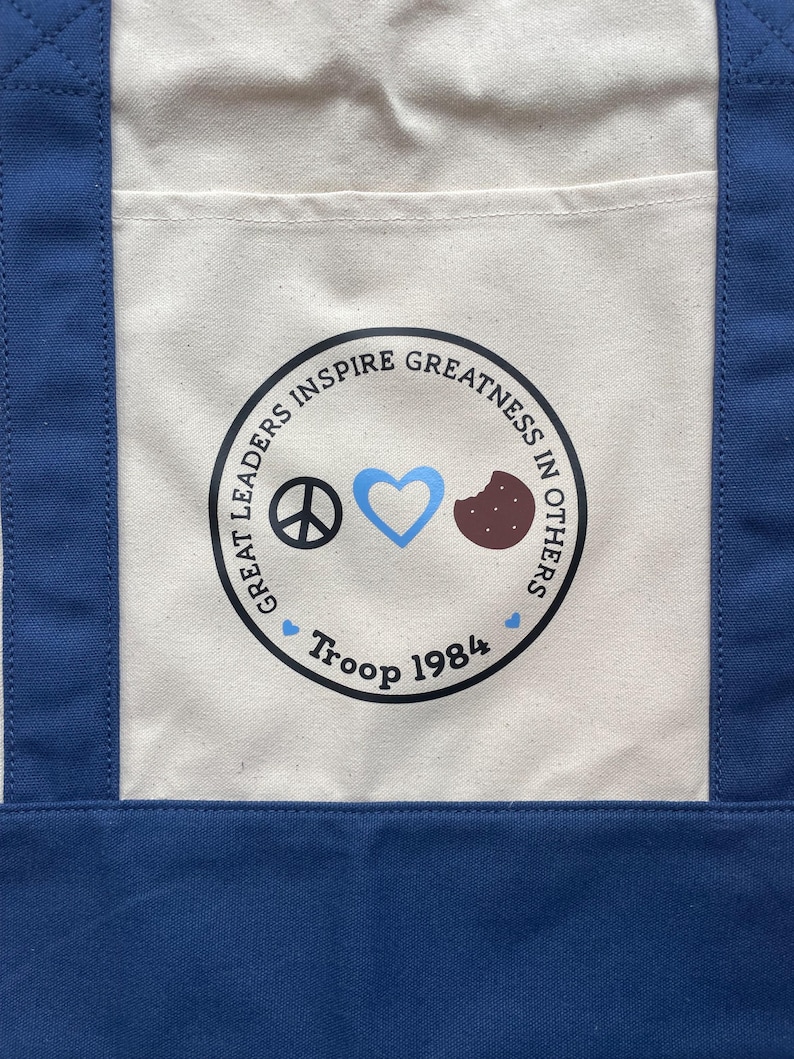 May include: A blue and cream tote bag with a circular graphic. The graphic includes a peace sign, a blue heart, and a cookie. The text reads "Great Leaders Inspire Greatness in Others" and "Troop 1984".