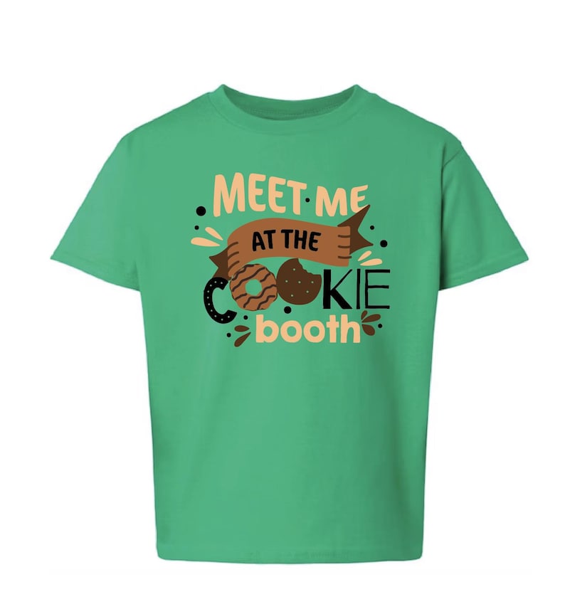 Puede incluir: Una camiseta verde con el texto "MEET ME AT THE COOKIE booth" en una fuente juguetona. El dise&ntilde;o incluye ilustraciones de galletas y una pancarta, ideal para un look casual.