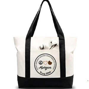 May include: A white canvas tote bag with black accents on the bottom and straps. The front pocket features a circular design with the words "GREAT LEADERS INSPIRE GREATNESS IN OTHERS," the name "Morgan," and "Troop 3960."