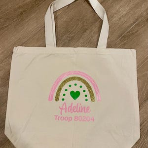 以下が含まれることがあります： ピンクとゴールドのグリッターの虹のデザイン、緑のハート、ピンクで「Adeline」と「Troop 80204」が描かれたクリーム色のトートバッグ。バッグにはクリーム色のハンドルが2つ付いています。