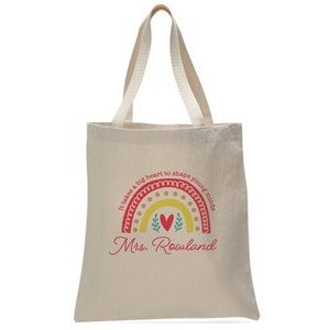 May include: A beige tote bag with a red and yellow rainbow design. The rainbow has a heart in the center and the text "It takes a big heart to shape young minds" around it. The text "Mrs. Rowland" is printed below the rainbow.