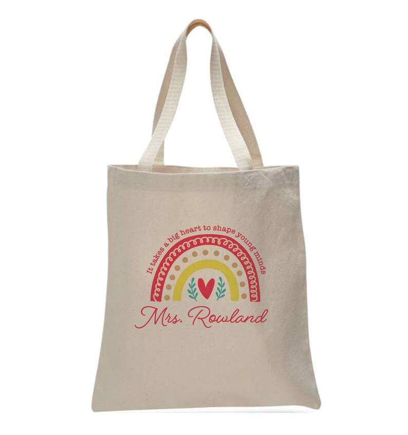 May include: A beige tote bag with a red and yellow rainbow design. The rainbow has a heart in the center and the text "It takes a big heart to shape young minds" around it. The text "Mrs. Rowland" is printed below the rainbow.