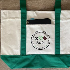 May include: Cream-colored tote bag with green accents. The bag features green handles and a green bottom. A front pocket displays a circular emblem with the text "GREAT LEADERS INSPIRE GREATNESS IN OTHERS" and the name "Jessica" and "Troop 684". A notebook is visible in the pocket.