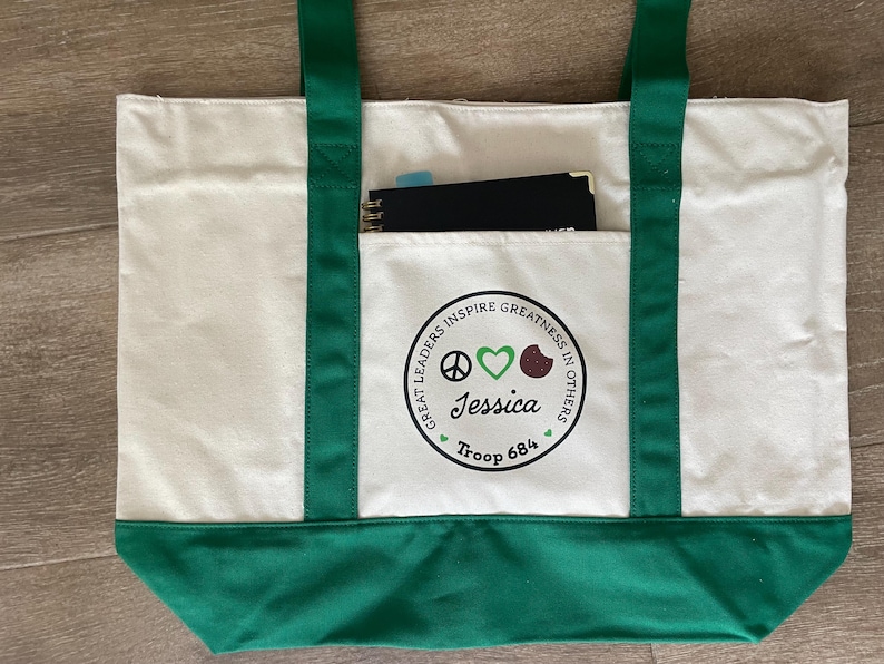 May include: Cream-colored tote bag with green accents. The bag features green handles and a green bottom. A front pocket displays a circular emblem with the text "GREAT LEADERS INSPIRE GREATNESS IN OTHERS" and the name "Jessica" and "Troop 684". A notebook is visible in the pocket.