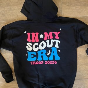 May include: Black hooded sweatshirt with a pink, white, and blue graphic that says "In My Scout Era" with a smiley face and a lightning bolt. The text "Troop 20226" is below the graphic.