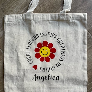 Pu&ograve; includere: Una borsa di tela bianca con testo nero che recita "Great Leaders Inspire Greatness in Others" con un fiore rosso con un viso sorridente giallo al centro. Il nome "Angelica" &egrave; scritto sotto il cerchio.