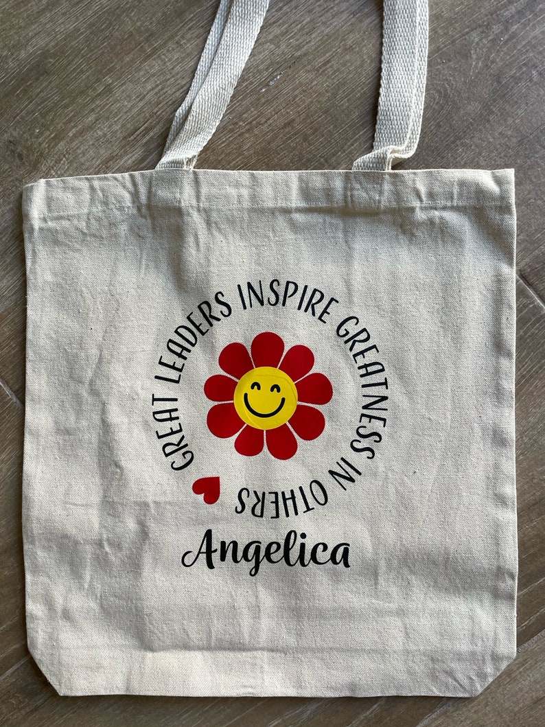 Pu&ograve; includere: Una borsa di tela bianca con testo nero che recita "Great Leaders Inspire Greatness in Others" con un fiore rosso con un viso sorridente giallo al centro. Il nome "Angelica" &egrave; scritto sotto il cerchio.