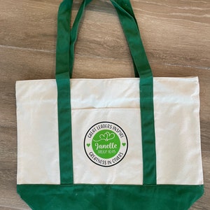 Pu&ograve; includere: Una borsa tote in tela bianca con manici e bordi verdi. La borsa ha un logo circolare con il testo "Great Leaders Inspire" e "Janelle Troop 1045" in un cerchio verde con due cuori. Il testo "Greatness in Others" &egrave; sotto il cerchio.