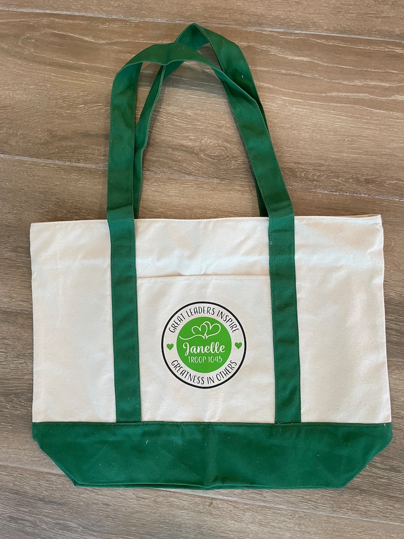 Pu&ograve; includere: Una borsa tote in tela bianca con manici e bordi verdi. La borsa ha un logo circolare con il testo "Great Leaders Inspire" e "Janelle Troop 1045" in un cerchio verde con due cuori. Il testo "Greatness in Others" &egrave; sotto il cerchio.