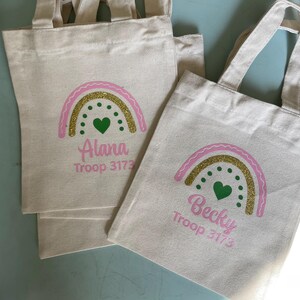 May include: Two off-white canvas tote bags with handles. Each bag features a printed graphic of a rainbow with a pink outline, a gold glitter arch, and green dots with a central green heart. The names Alana and Becky are printed in pink.