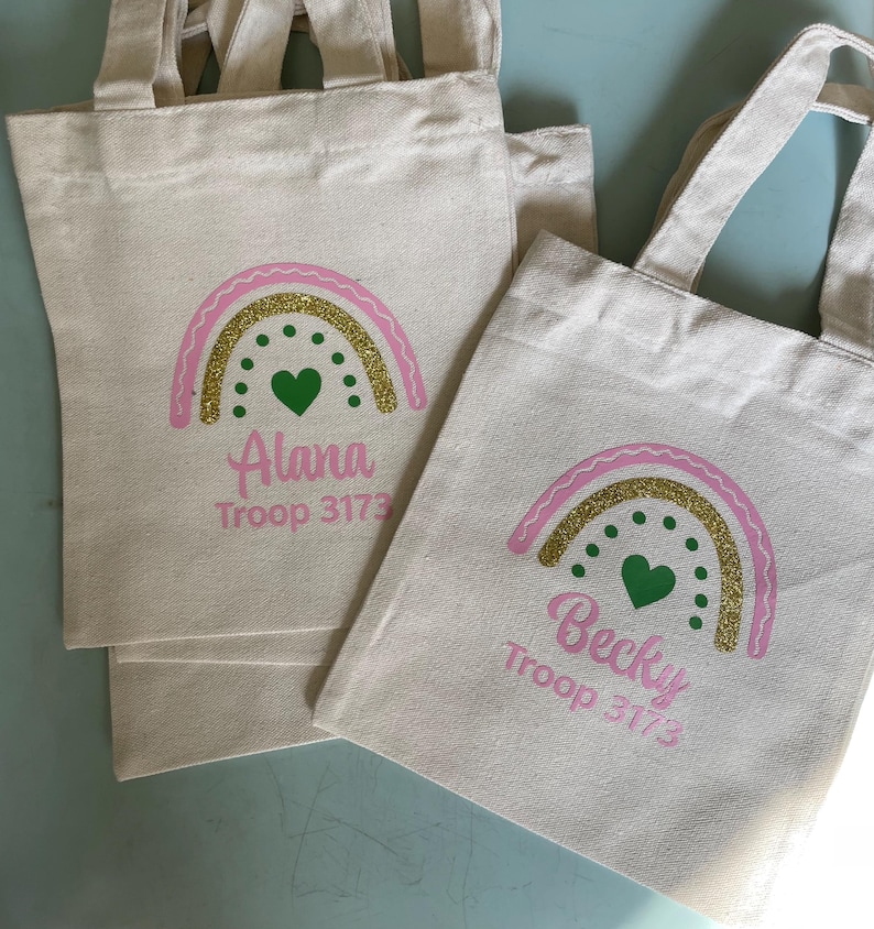 May include: Two off-white canvas tote bags with handles. Each bag features a printed graphic of a rainbow with a pink outline, a gold glitter arch, and green dots with a central green heart. The names Alana and Becky are printed in pink.