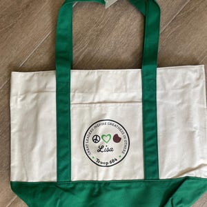 May include: A white canvas tote bag with green accents. The bag features green handles and a green bottom panel. A circular emblem on the front includes the text "Great Leaders Inspire Greatness in Others," a peace sign, a heart, and the name "Lisa" with "Troop 684."