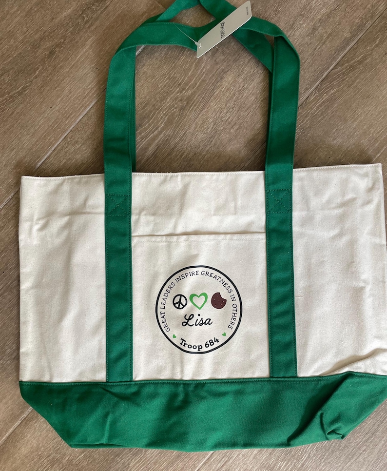 May include: A white canvas tote bag with green accents. The bag features green handles and a green bottom panel. A circular emblem on the front includes the text "Great Leaders Inspire Greatness in Others," a peace sign, a heart, and the name "Lisa" with "Troop 684."