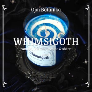 May include: A blue glass candle with a white and blue swirl design. The candle has a label that reads "Whimsigoth" and "vanilla, sea salt, jasmine & sheer". The candle is on a decorative silver tray. The text "Ojai Botanika" is at the top.