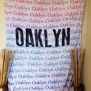 May include: A white fabric banner with the name "Oaklyn" printed in black letters. The banner is decorated with a ruffled edge and has the name "Oaklyn" repeated multiple times in various colors.