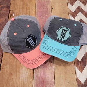 May include: Two trucker hats with a black and white graphic of a racing flag on the front. One hat is gray with a coral brim and the other is gray with a turquoise brim.