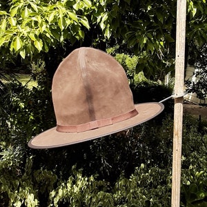 May include: A brown leather hat with a wide brim hanging on a wooden pole. The hat has a tall, pointed crown and a brown ribbon around the base.