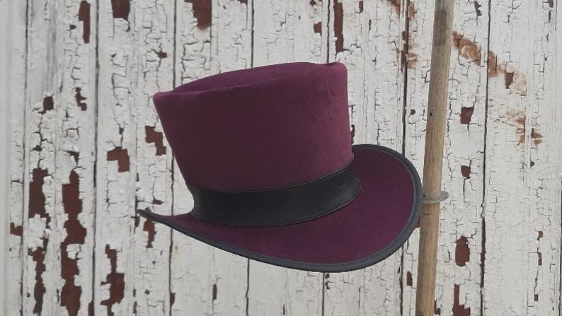 May include: A burgundy top hat with a black band. The hat is made of felt and has a wide brim.