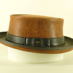 Puede incluir: Un sombrero fedora de cuero marrón con una banda de cuero negro y una hebilla dorada. El sombrero tiene un ala ancha y una copa alta.