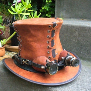 May include: A brown leather top hat with a black leather band and brass eyepieces. The hat has a lace-up detail on the side.