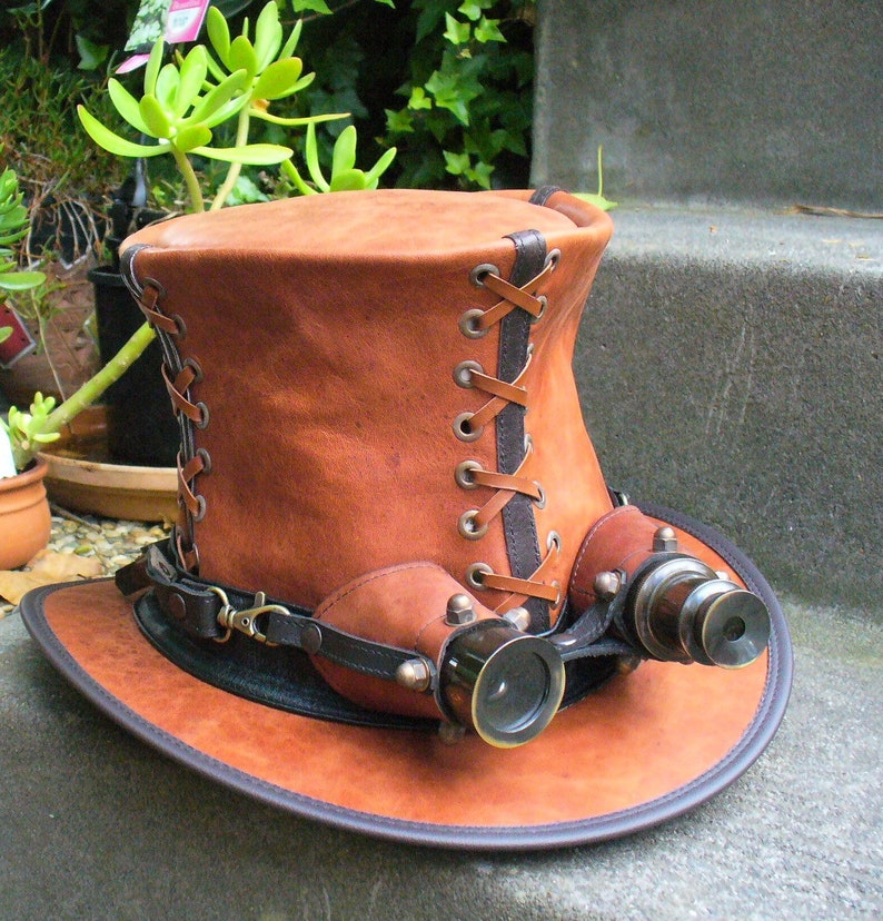 May include: A brown leather top hat with a black leather band and brass eyepieces. The hat has a lace-up detail on the side.