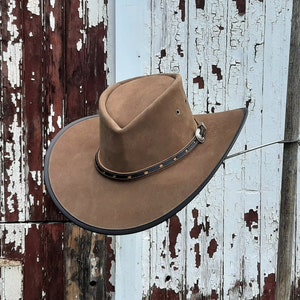 May include: A brown leather cowboy hat with a black band and a silver buckle. The hat is hanging on a wooden post against a weathered wooden background.