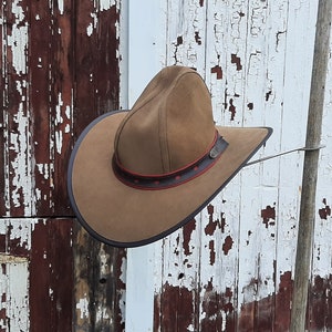 May include: A brown leather cowboy hat with a red and black band hanging on a wooden post against a white and brown weathered wooden background.