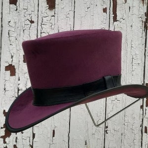 May include: A purple top hat with a black band around the crown. The hat is displayed on a stand against a white wooden background.