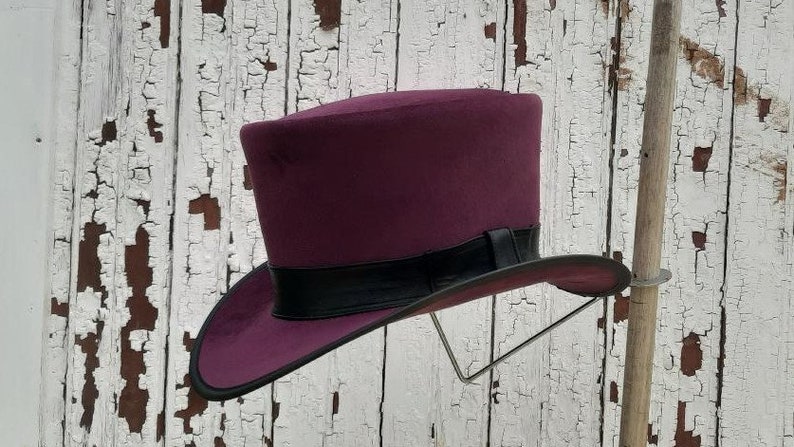May include: A purple top hat with a black band around the crown. The hat is displayed on a stand against a white wooden background.
