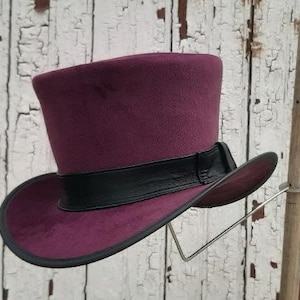 May include: A burgundy top hat with a black leather band. The hat is displayed on a wooden stand against a white wooden background.